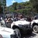 A cluster of people sit in 2 seater buggies in a parking lot with a backdrop of trees near a sign that reads Buggy VTT Rallye Adventure